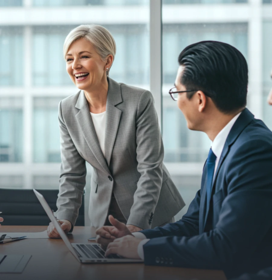 Smiling businesswoman in a gray suit leading a meeting with colleagues seated around a conference table in a modern office.