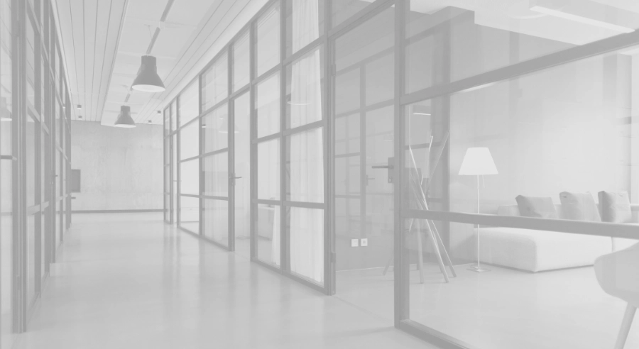 Modern office hallway with glass-walled meeting rooms, pendant lighting, and a lounge area with a sofa and floor lamp.
